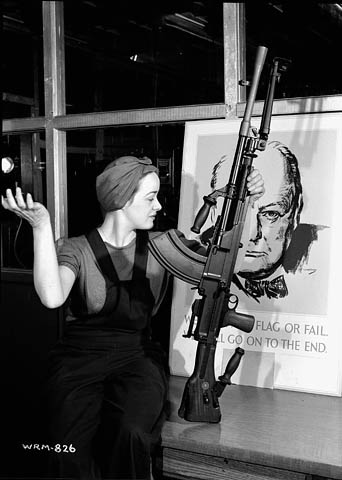 À l’usine de fusils-mitrailleurs Bren de la John Inglis Co., Veronica (Ronnie) Foster, employée de cette société et connue sous le nom de « la fille au fusil-mitrailleur », pose avec un de ces fusils terminé devant une affiche de Winston Churchill